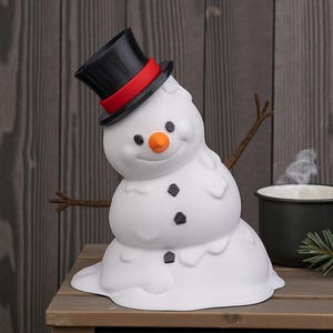 May include: A novelty white snowman ornament with a black top hat and red band, orange nose, and black button features. The snowman has twig arms and a melting design. It sits on a wooden surface.
