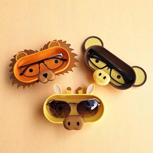 Puede incluir: Tres sujetagafas con tem&aacute;tica animal en naranja, marr&oacute;n y amarillo. Los dise&ntilde;os de le&oacute;n, mono y jirafa presentan cada uno una bandeja para guardar las gafas, con las monturas visibles. Los soportes est&aacute;n sobre un fondo naranja claro.