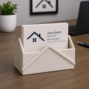 Pu&ograve; includere: Un portacarte beige a forma di casa, contenente una pila di biglietti da visita bianchi. Le carte mostrano un logo di casa blu scuro e il nome "Jane Smith, REALTOR&reg;" con informazioni di contatto. Una piccola pianta e un logo di casa incorniciato sono sullo sfondo.