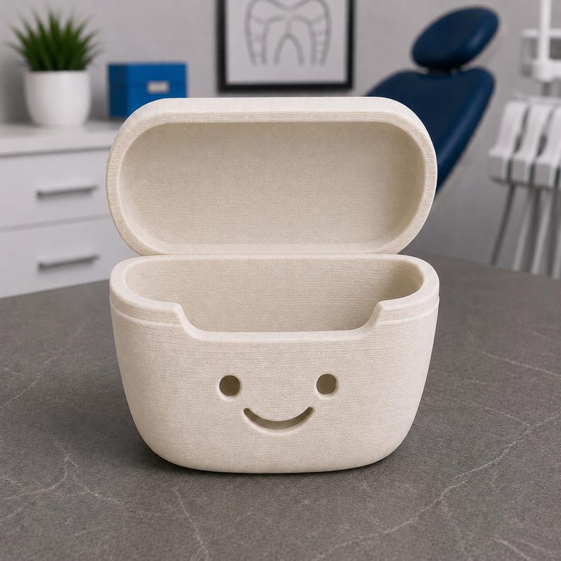 May include: An open, beige-colored case with a smiling face design. The case has a rectangular shape with rounded edges and a textured surface. The background includes a dental chair and a framed dental illustration.