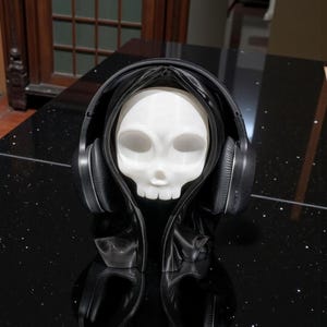 May include: A white skull figure wearing a black hooded cloak and black headphones. The skull has large eye sockets and a small nose. The headphones are over the ears. The figure is on a black surface.