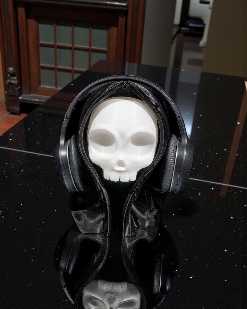 May include: A white skull figure wearing a black hooded cloak and black headphones. The skull has large eye sockets and a small nose. The headphones are over the ears. The figure is on a black surface.