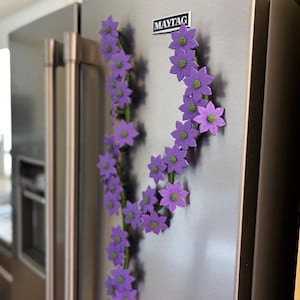 May include: A decorative floral magnet arrangement on a stainless steel refrigerator. The arrangement features purple flowers with green centers, arranged in a curved pattern. The refrigerator has a "MAYTAG" logo.