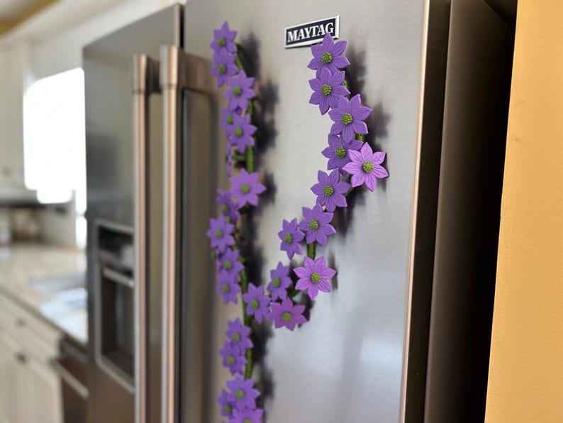 May include: A decorative floral magnet arrangement on a stainless steel refrigerator. The arrangement features purple flowers with green centers, arranged in a curved pattern. The refrigerator has a "MAYTAG" logo.