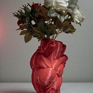 May include: A red, heart-shaped vase holding a bouquet of red and white roses with green leaves. The vase has a glossy finish and is a unique decorative piece.