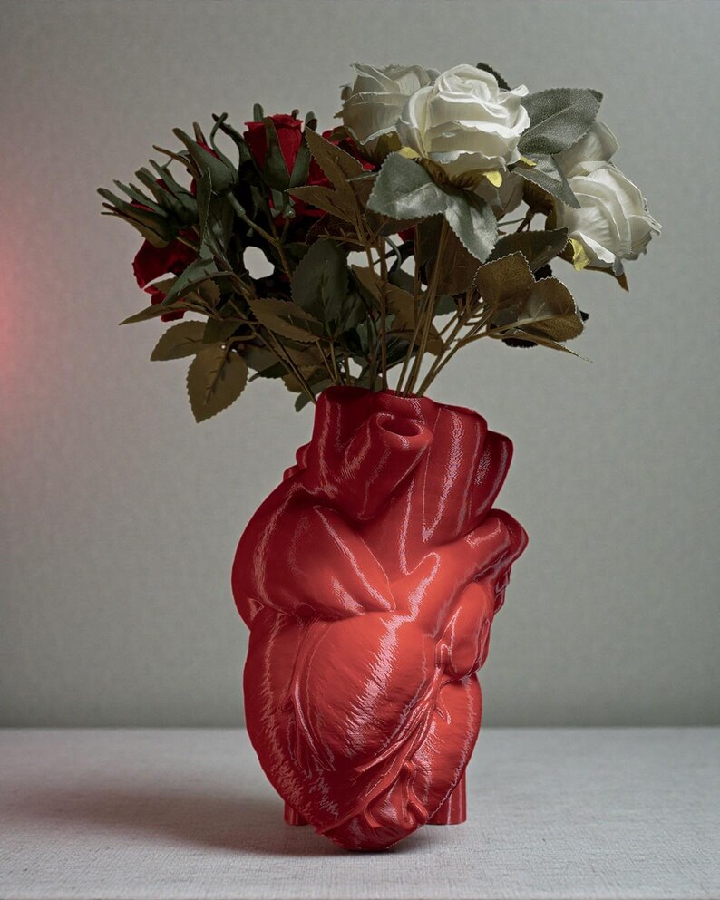 May include: A red, heart-shaped vase holding a bouquet of red and white roses with green leaves. The vase has a glossy finish and is a unique decorative piece.