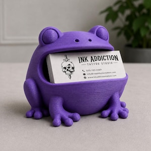 May include: A purple frog-shaped business card holder. The frog has large eyes and a wide mouth holding a stack of white business cards. The cards display the text "INK ADDICTION - TATTOO STUDIO" along with contact information.