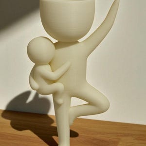 Peut inclure: Figurine imprimée en 3D de couleur crème représentant une personne tenant une plus petite figurine. La plus grande figure a un bras levé et une tête en forme de bol, tandis que la plus petite est bercée dans ses bras. La figurine est sur une surface en bois.