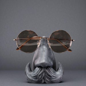 May include: A novelty eyeglass holder shaped like a face with a mustache and nose, holding a pair of round, gold-rimmed sunglasses. The holder is gray and the sunglasses have brown lenses.