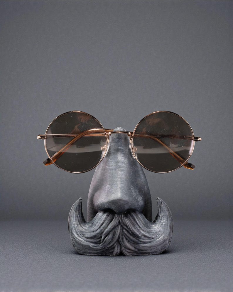 May include: A novelty eyeglass holder shaped like a face with a mustache and nose, holding a pair of round, gold-rimmed sunglasses. The holder is gray and the sunglasses have brown lenses.