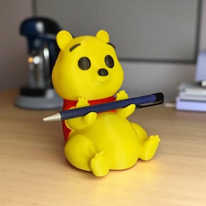 May include: A yellow Winnie the Pooh figurine holding a blue pen. The figurine has black eyes and a red shirt. The pen is held in a writing position, suggesting it is a pen holder or desk accessory.