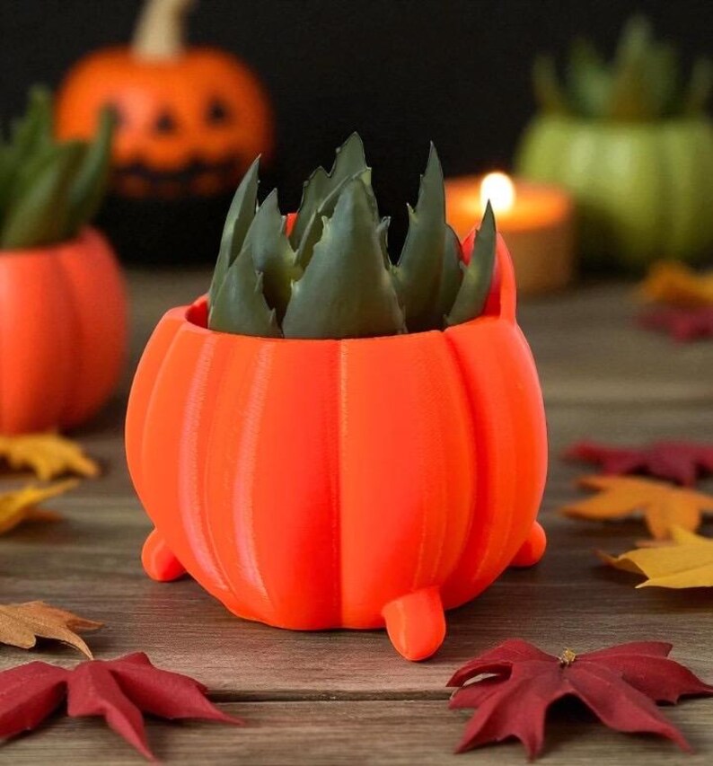 May include: An orange pumpkin-shaped planter with a green succulent plant inside. The planter has three small feet and vertical ridges. Other pumpkin planters and fall leaves are in the background, creating a festive autumn scene.