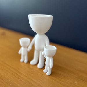 May include: Three white ceramic figurines in a family arrangement. The largest figure stands in the center, holding the hand of a smaller figure on the left. A third, smaller figure is on the right, with an arm raised. The figures have a minimalist design.