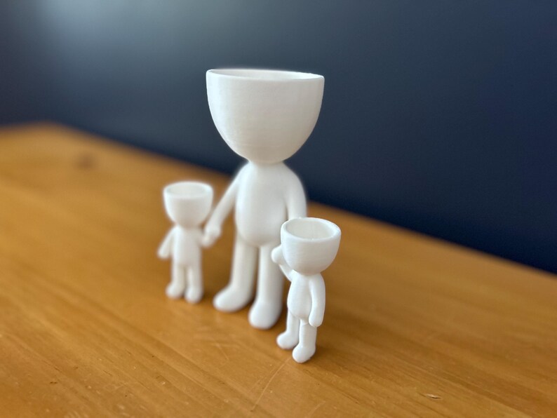 May include: Three white ceramic figurines in a family arrangement. The largest figure stands in the center, holding the hand of a smaller figure on the left. A third, smaller figure is on the right, with an arm raised. The figures have a minimalist design.
