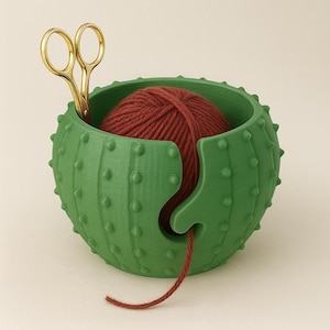 May include: A green cactus-shaped yarn bowl with a red yarn ball inside. Gold scissors are placed in the bowl. The bowl has a slot to feed the yarn through. The bowl is designed to keep yarn clean and prevent it from rolling away.