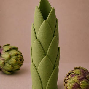 May include: A green, artichoke-shaped vase with a textured surface. The vase is surrounded by two artichokes with green and purple hues. The vase is a decorative item, perfect for adding a touch of nature-inspired design to any space.