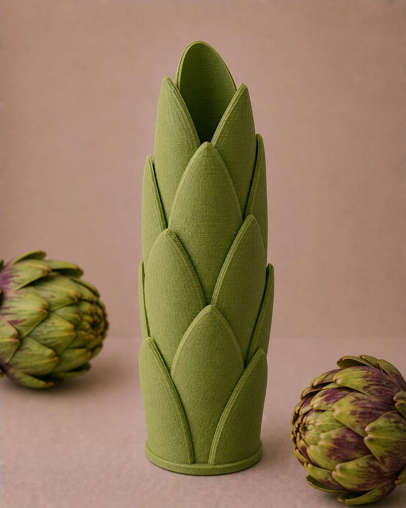 May include: A green, artichoke-shaped vase with a textured surface. The vase is surrounded by two artichokes with green and purple hues. The vase is a decorative item, perfect for adding a touch of nature-inspired design to any space.
