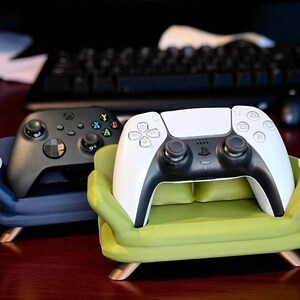 May include: A black Xbox controller and a white and black PlayStation controller rest on miniature sofa-shaped stands. The green and blue stands have wooden legs. The controllers are set against a blurred background.