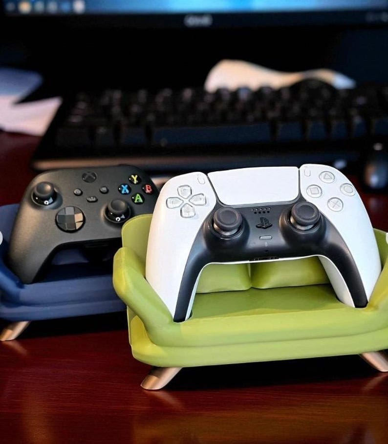 May include: A black Xbox controller and a white and black PlayStation controller rest on miniature sofa-shaped stands. The green and blue stands have wooden legs. The controllers are set against a blurred background.