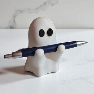 May include: A white ghost-shaped pen holder with black eyes, holding a dark blue pen with a silver tip. The pen holder is made of a smooth material and sits on a white surface. The pen is held horizontally.