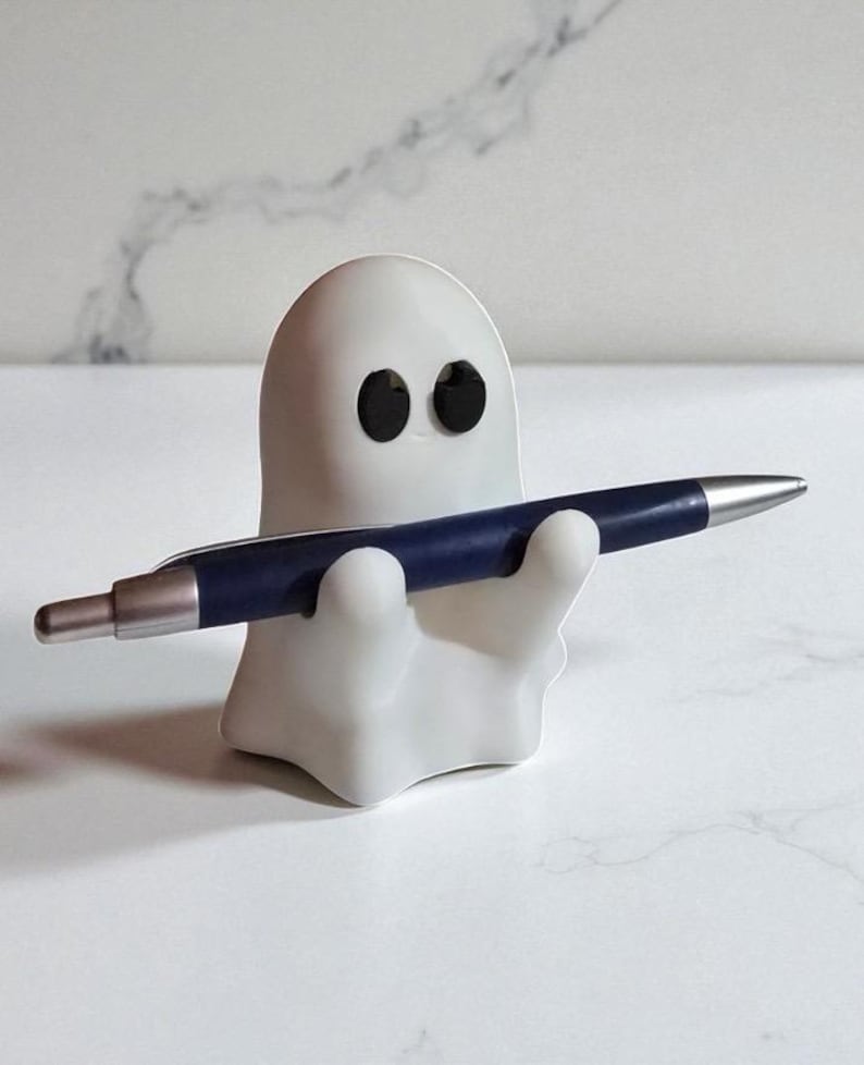 May include: A white ghost-shaped pen holder with black eyes, holding a dark blue pen with a silver tip. The pen holder is made of a smooth material and sits on a white surface. The pen is held horizontally.