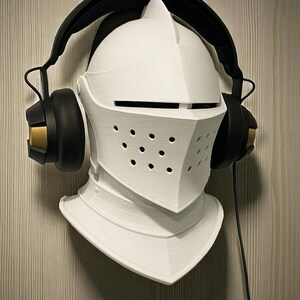 May include: A white, 3D-printed knight's helmet with a black headset. The helmet has a visor with small holes and is mounted on a wall. The headset has black earcups with gold accents and a black headband.