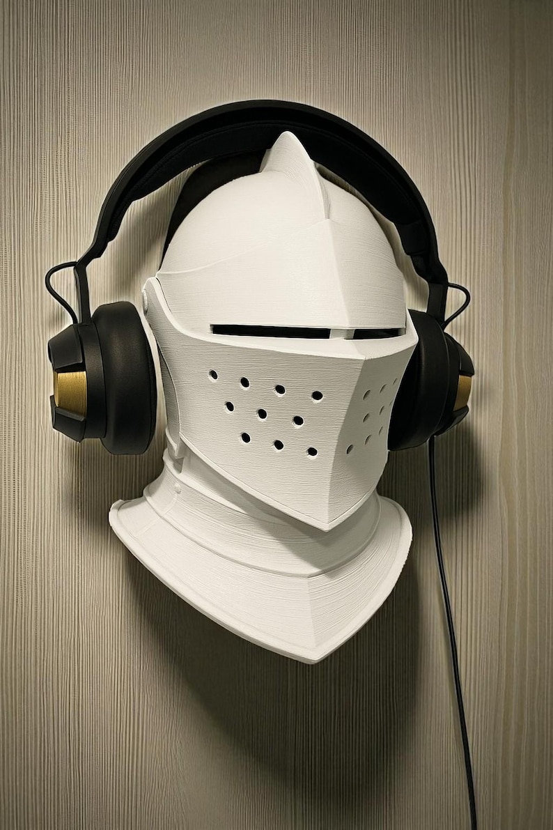 May include: A white, 3D-printed knight's helmet with a black headset. The helmet has a visor with small holes and is mounted on a wall. The headset has black earcups with gold accents and a black headband.
