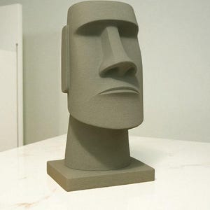 May include: A gray Moai statue replica, featuring a large rectangular head with a prominent nose and a stern expression. The statue is set on a square base, showcasing a textured surface. The sculpture is a decorative item.