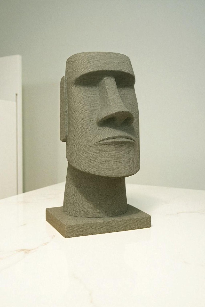May include: A gray Moai statue replica, featuring a large rectangular head with a prominent nose and a stern expression. The statue is set on a square base, showcasing a textured surface. The sculpture is a decorative item.