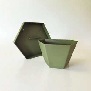 May include: An olive green, geometric planter with a matching hexagonal back panel. The planter has a tapered shape and is open at the top. The back panel has two holes for mounting and a small handle. The items are made of metal.
