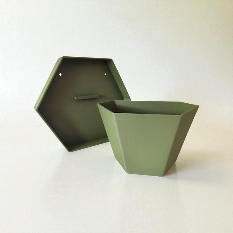 May include: An olive green, geometric planter with a matching hexagonal back panel. The planter has a tapered shape and is open at the top. The back panel has two holes for mounting and a small handle. The items are made of metal.