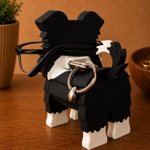 May include: A black and white dog-shaped eyeglasses and ring holder. The holder has a black body with white accents and feet. Eyeglasses are placed on the dog's head, and a ring is on the tail. The holder is on a wooden surface.