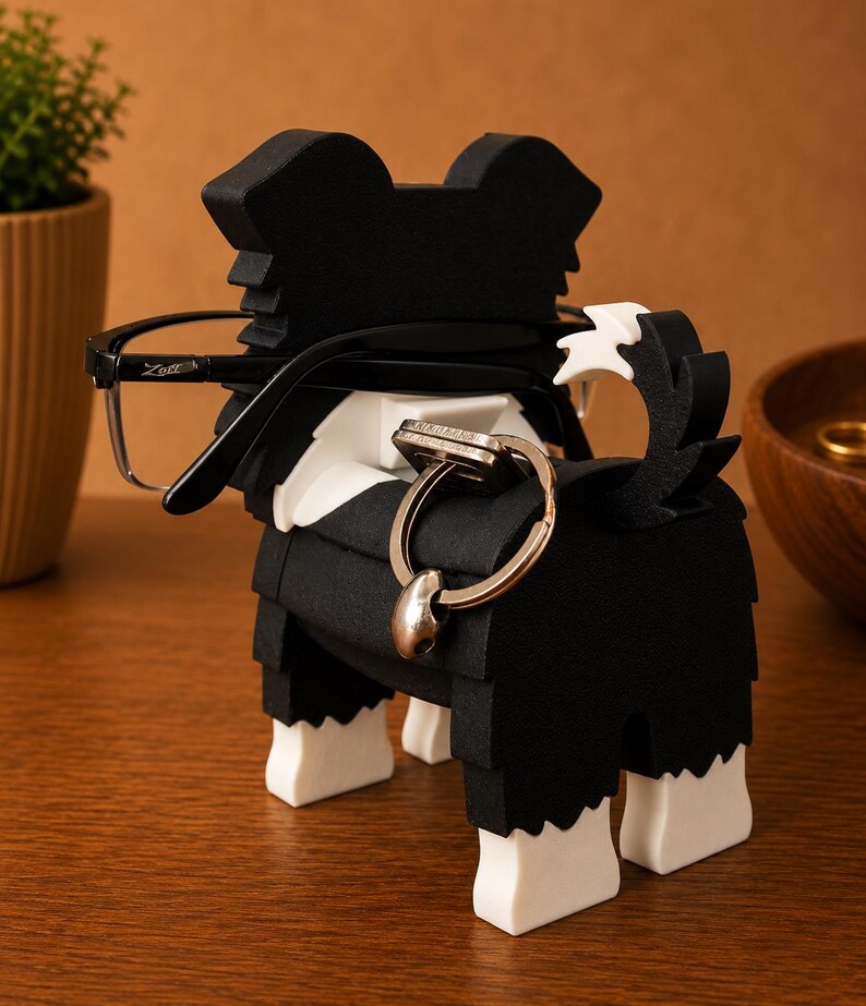 May include: A black and white dog-shaped eyeglasses and ring holder. The holder has a black body with white accents and feet. Eyeglasses are placed on the dog's head, and a ring is on the tail. The holder is on a wooden surface.