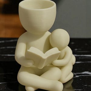 May include: A cream-colored figurine depicting a parent figure reading a book to a child. The parent is seated with legs crossed, holding the book open. The child is nestled close, embracing the parent. The top of the parent figure is a planter.