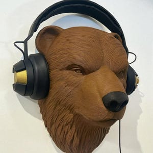 Puede incluir: Escultura de una cabeza de oso pardo con auriculares negros y dorados. El oso tiene una nariz negra y est&aacute; montado en una pared blanca. Los auriculares tienen un cable negro colgando.