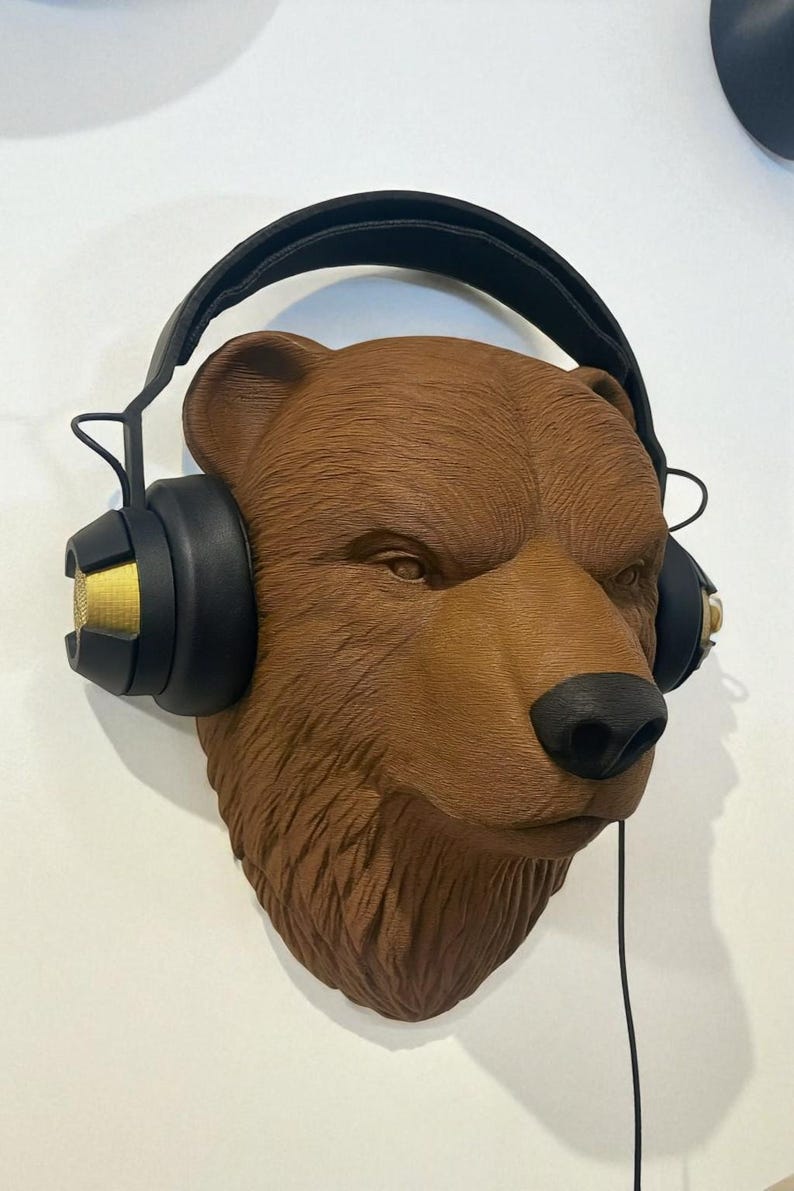Puede incluir: Escultura de una cabeza de oso pardo con auriculares negros y dorados. El oso tiene una nariz negra y est&aacute; montado en una pared blanca. Los auriculares tienen un cable negro colgando.