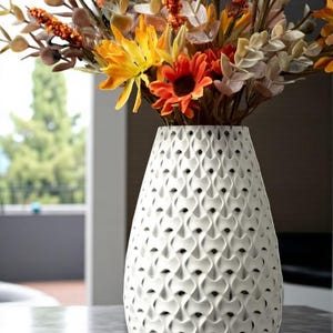 May include: A white ceramic vase with a textured, repeating pattern holds a vibrant arrangement of artificial flowers. The flowers feature orange, yellow, and brown hues, creating a warm, autumnal aesthetic. The vase is on a gray surface.