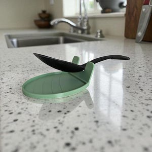 May include: A sage green silicone spoon rest with a leaf design, holding a black spoon. The spoon rest is on a speckled countertop, providing a convenient and stylish place to set utensils while cooking.