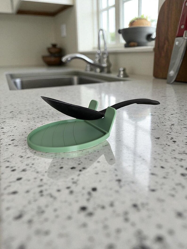 May include: A sage green silicone spoon rest with a leaf design, holding a black spoon. The spoon rest is on a speckled countertop, providing a convenient and stylish place to set utensils while cooking.