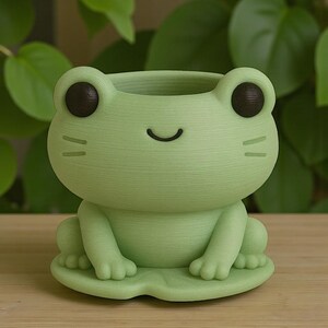 Puede incluir: Una maceta con forma de rana verde claro con una cara sonriente, ojos negros y dos peque&ntilde;as l&iacute;neas a cada lado de la cara. La maceta se asienta sobre una base en forma de hoja y est&aacute; hecha de un material texturizado. La maceta de rana es un art&iacute;culo decorativo.