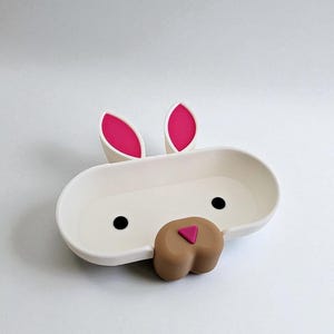May include: A white bunny-shaped soap dish with pink inner ears, black eyes, and a brown nose. The dish is oval-shaped with a shallow basin. The design is whimsical and suitable for a child's bathroom.