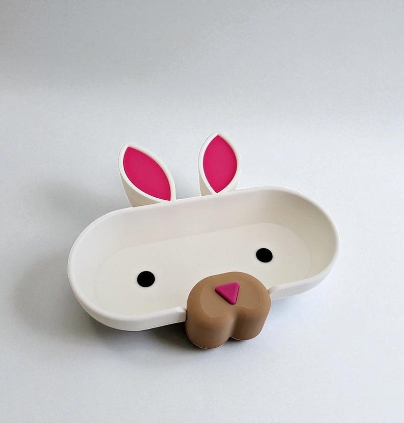 May include: A white bunny-shaped soap dish with pink inner ears, black eyes, and a brown nose. The dish is oval-shaped with a shallow basin. The design is whimsical and suitable for a child's bathroom.