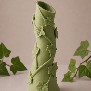 May include: A green vase with a textured surface and a curved shape. The vase is adorned with ivy vines and leaves. The vase is approximately 10 inches tall.