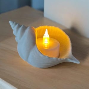 Può includere: Un portacandele a forma di conchiglia bianca con una candela LED accesa all'interno. Il portacandele è in ceramica e ha una finitura liscia e opaca. La candela emette una luce gialla calda, creando un'atmosfera accogliente. Su una superficie di legno.