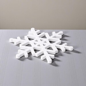 Puede incluir: Un objeto decorativo blanco con forma de copo de nieve. El copo de nieve tiene un dise&ntilde;o calado detallado con seis puntas sim&eacute;tricas. Est&aacute; hecho de un material s&oacute;lido y opaco y se encuentra sobre una superficie gris. Decoraci&oacute;n con tem&aacute;tica invernal.