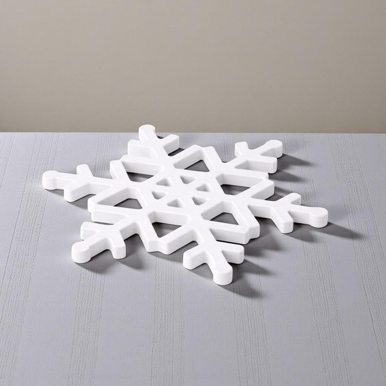 Puede incluir: Un objeto decorativo blanco con forma de copo de nieve. El copo de nieve tiene un dise&ntilde;o calado detallado con seis puntas sim&eacute;tricas. Est&aacute; hecho de un material s&oacute;lido y opaco y se encuentra sobre una superficie gris. Decoraci&oacute;n con tem&aacute;tica invernal.
