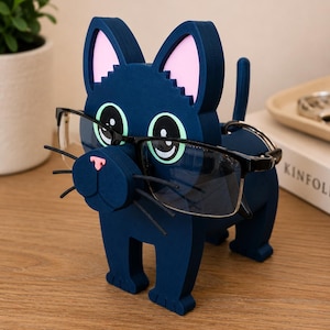 May include: A navy blue cat-shaped glasses holder with pink inner ears and green eyes. The holder is made of a solid material and is holding a pair of black-framed glasses. The cat has a pink nose and whiskers.