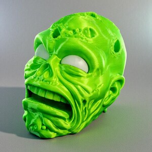 May include: A 3D-printed, lime green zombie head with a gaping mouth and white eyes. The head has a textured surface with visible print lines and several holes. The design is a grotesque, yet detailed, representation of a zombie.