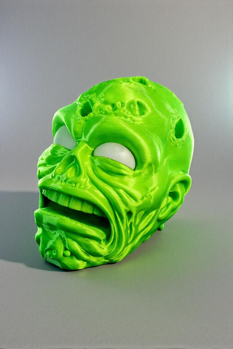 May include: A 3D-printed, lime green zombie head with a gaping mouth and white eyes. The head has a textured surface with visible print lines and several holes. The design is a grotesque, yet detailed, representation of a zombie.