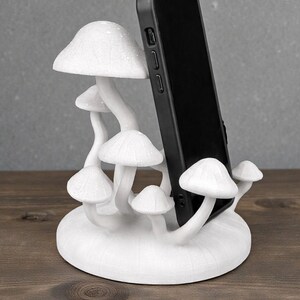 May include: A white mushroom-shaped phone stand with a black phone in place. The stand features multiple mushroom caps and stems, creating a whimsical and decorative design. The stand is made of a solid material and sits on a wooden surface.